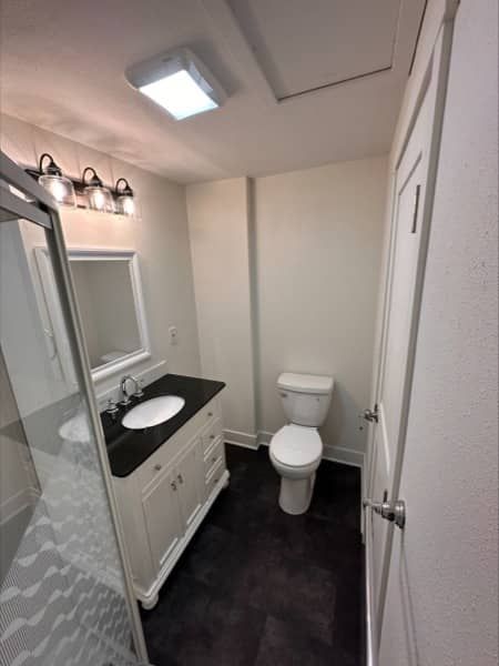 Bathroom with a vanity, toilet, and shower. White walls, dark floor.