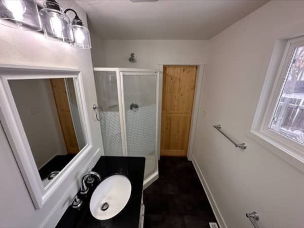 Bathroom with shower, vanity, wood door, and a window; white walls, black countertop, and brown floor.