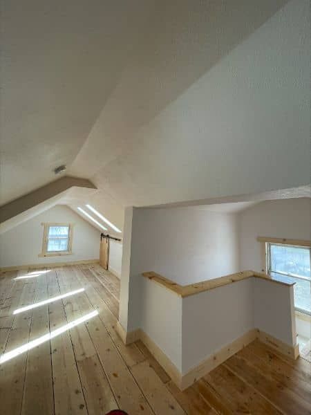 Bright loft interior with wood floor, sloped ceiling, small windows, and a railing.