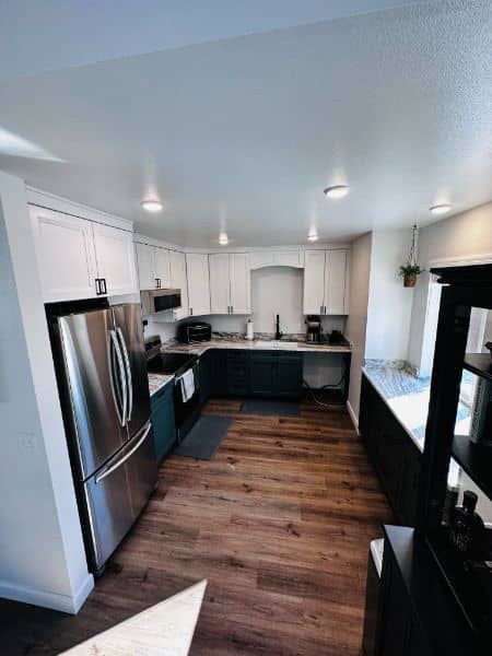 Modern kitchen with white upper cabinets, dark teal lower cabinets, stainless steel fridge, and wood-look flooring.