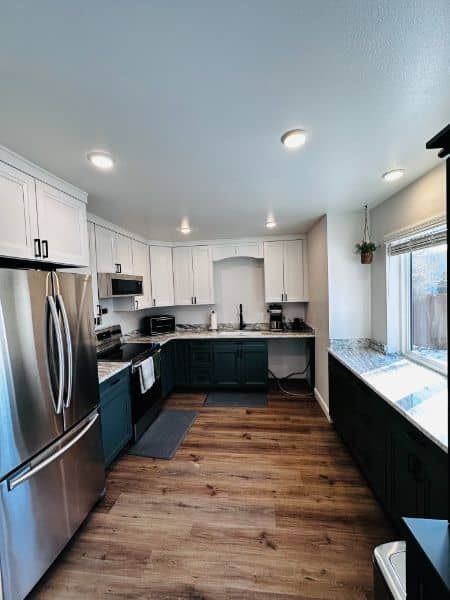 A kitchen with white upper cabinets, teal lower cabinets, stainless steel appliances, and wood-look flooring.