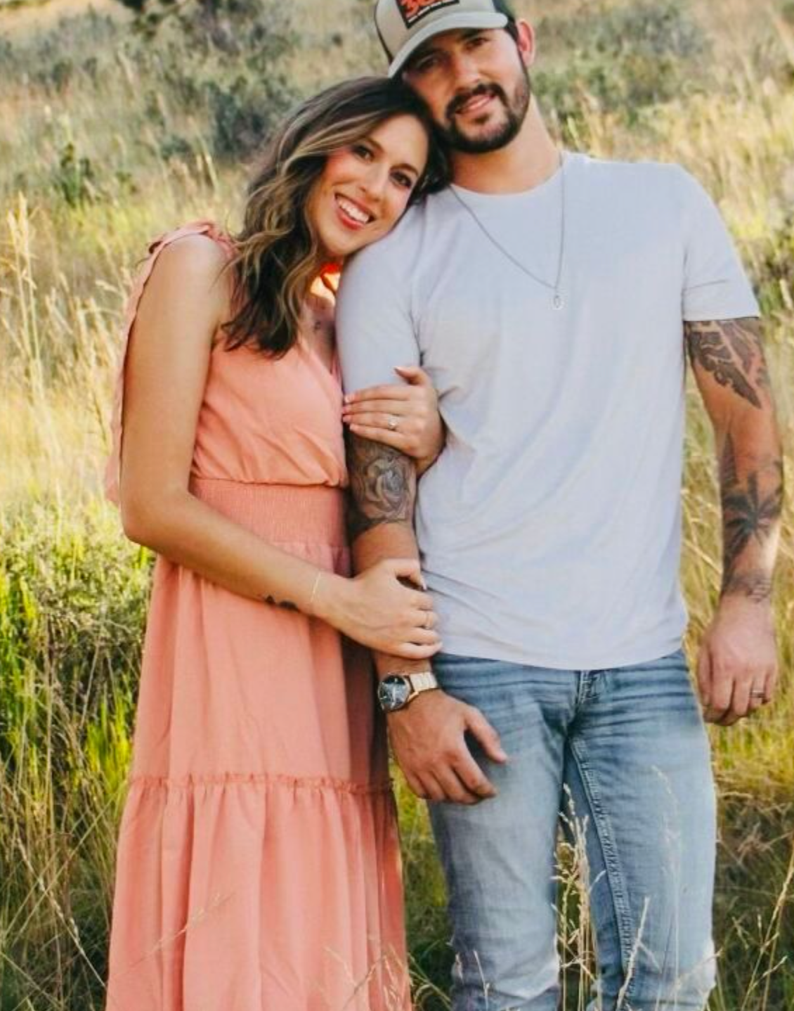 Woman in peach dress leans on man in white tee and jeans in field.