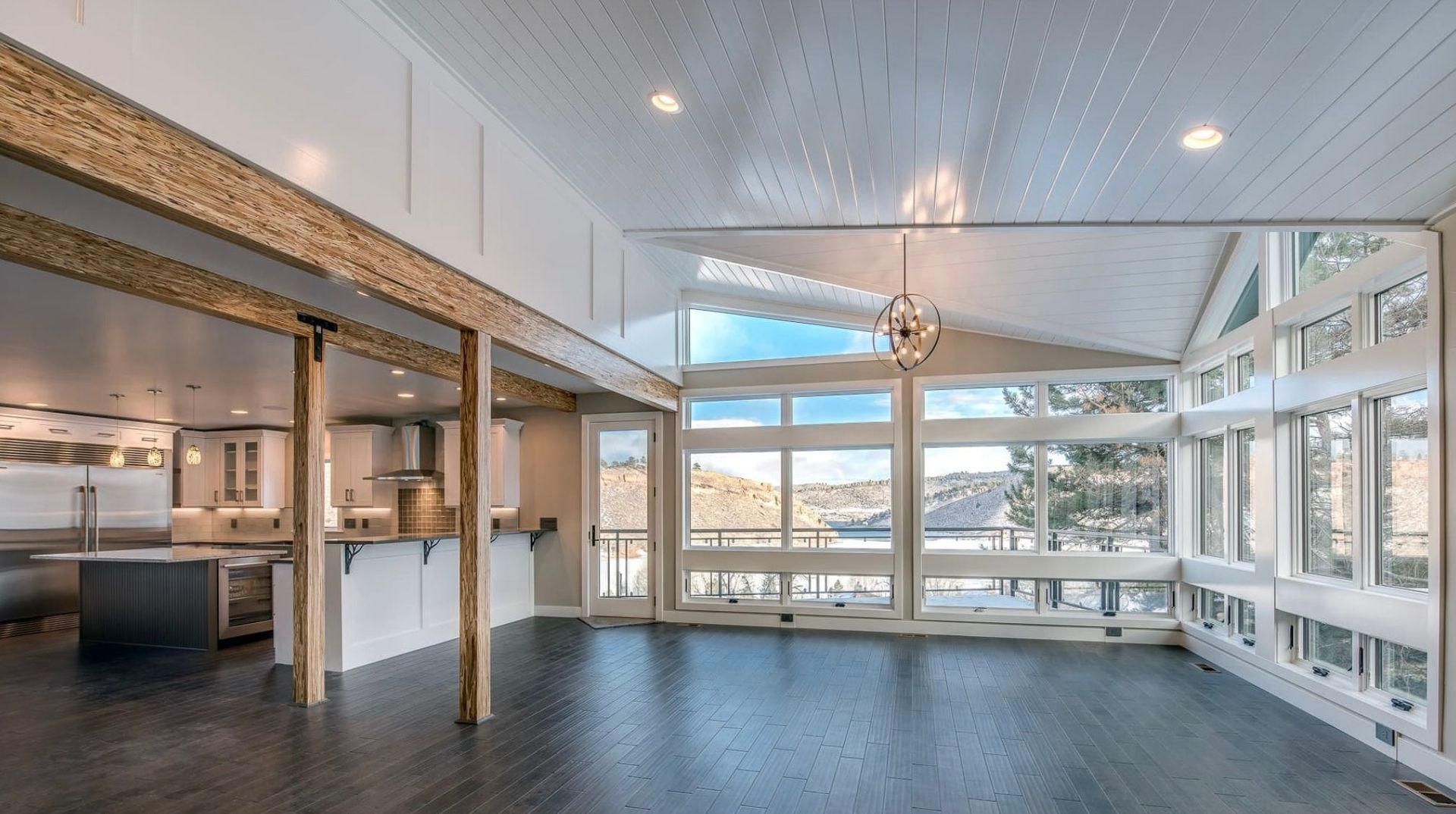 Open-concept living space with dark wood floors, large windows overlooking a snowy landscape, and exposed wood beams.