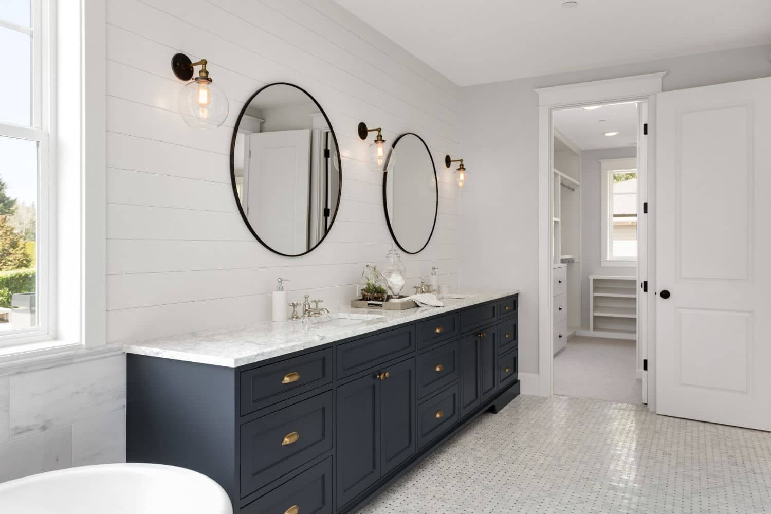 Bathroom with navy vanity, oval mirrors, white shiplap wall, and doorway to a closet.