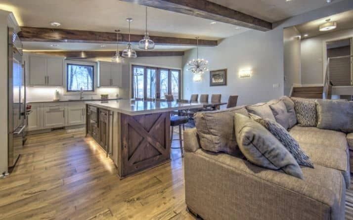Open-plan kitchen and living area with a large island, sofa, and dining table; rustic beams, hardwood floors.