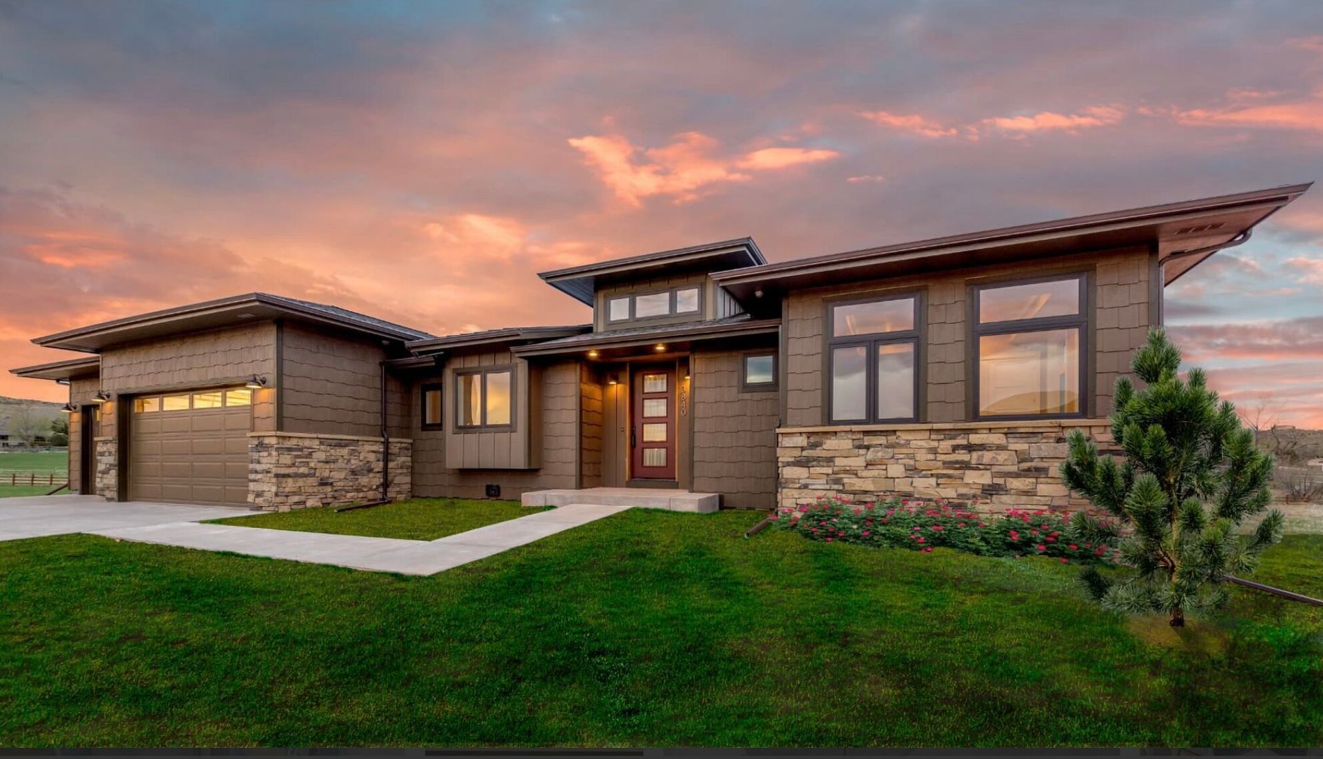 Modern brown house with stone accents, green lawn, and a sunset sky.