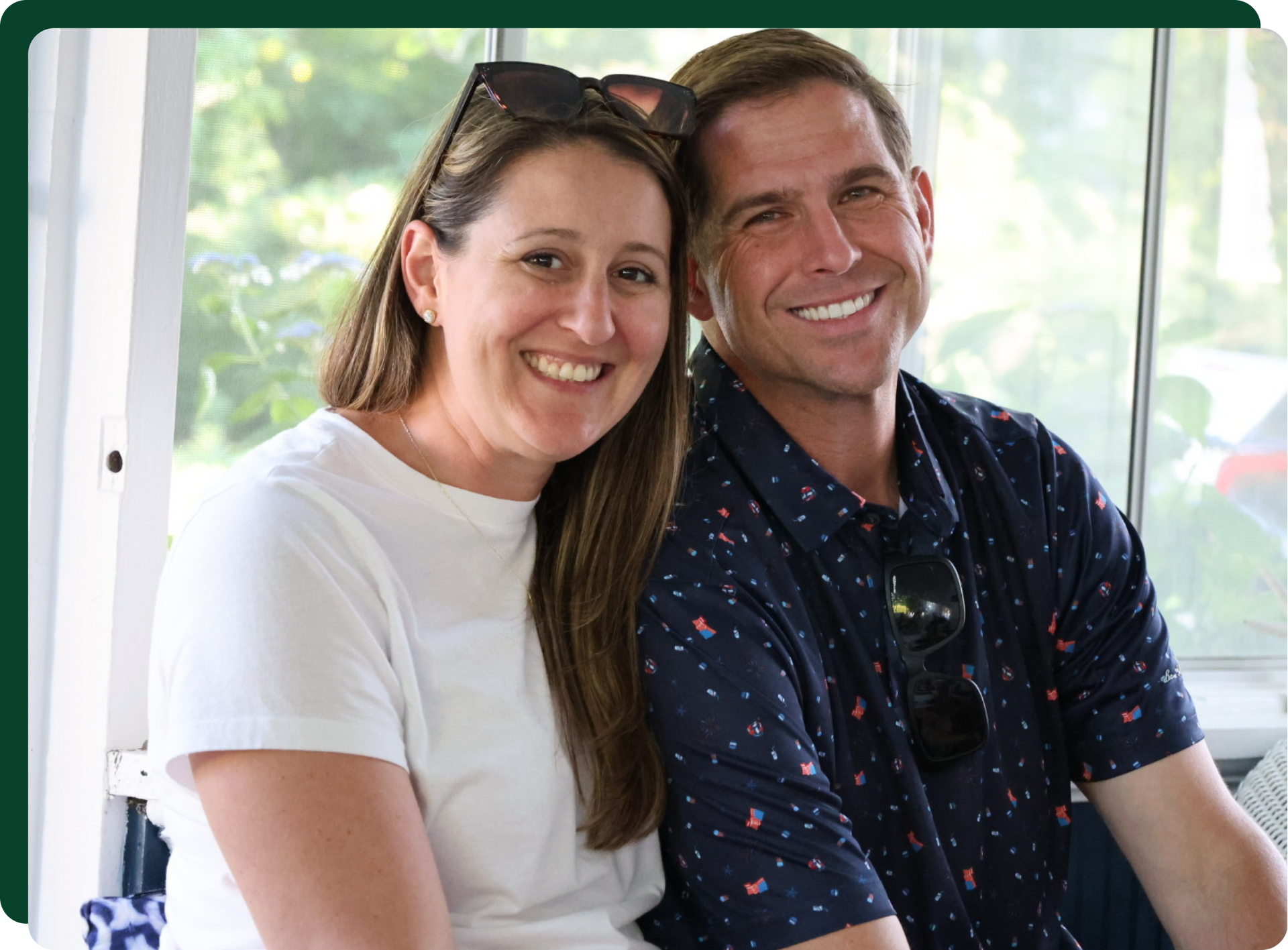 Smiling couple; woman in white shirt, man in a patterned shirt, both outdoors near a window.