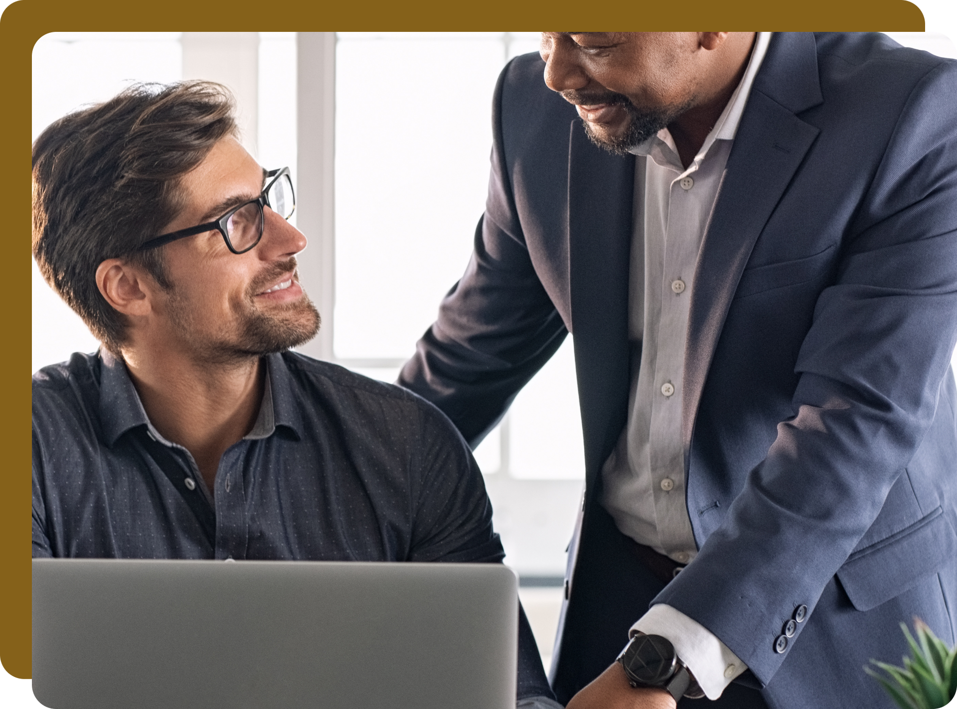 Two men smiling, looking at each other. One is sitting at a laptop, the other is leaning in, both in a modern office.