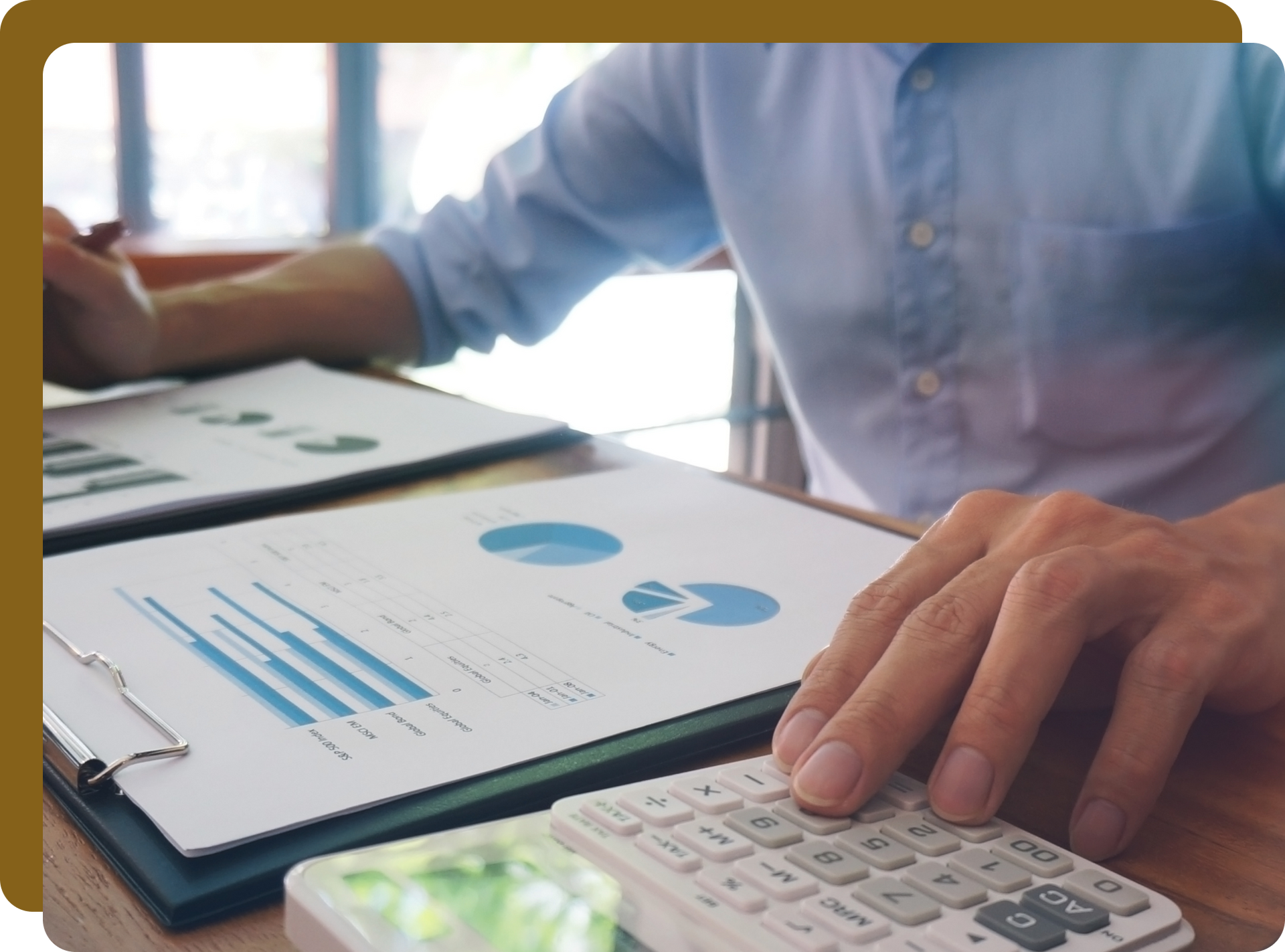 Person using a calculator, examining charts and graphs at a desk, in a light-filled office.