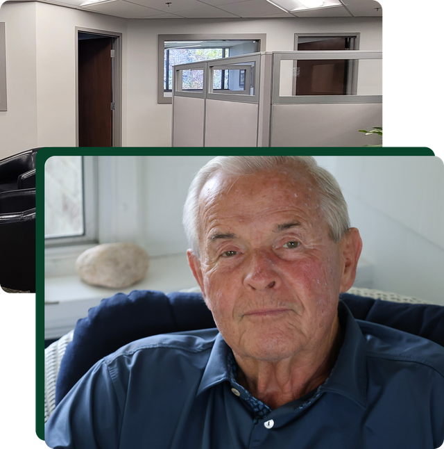 Senior man in a blue shirt sits in front of a window; office cubicles in the background.