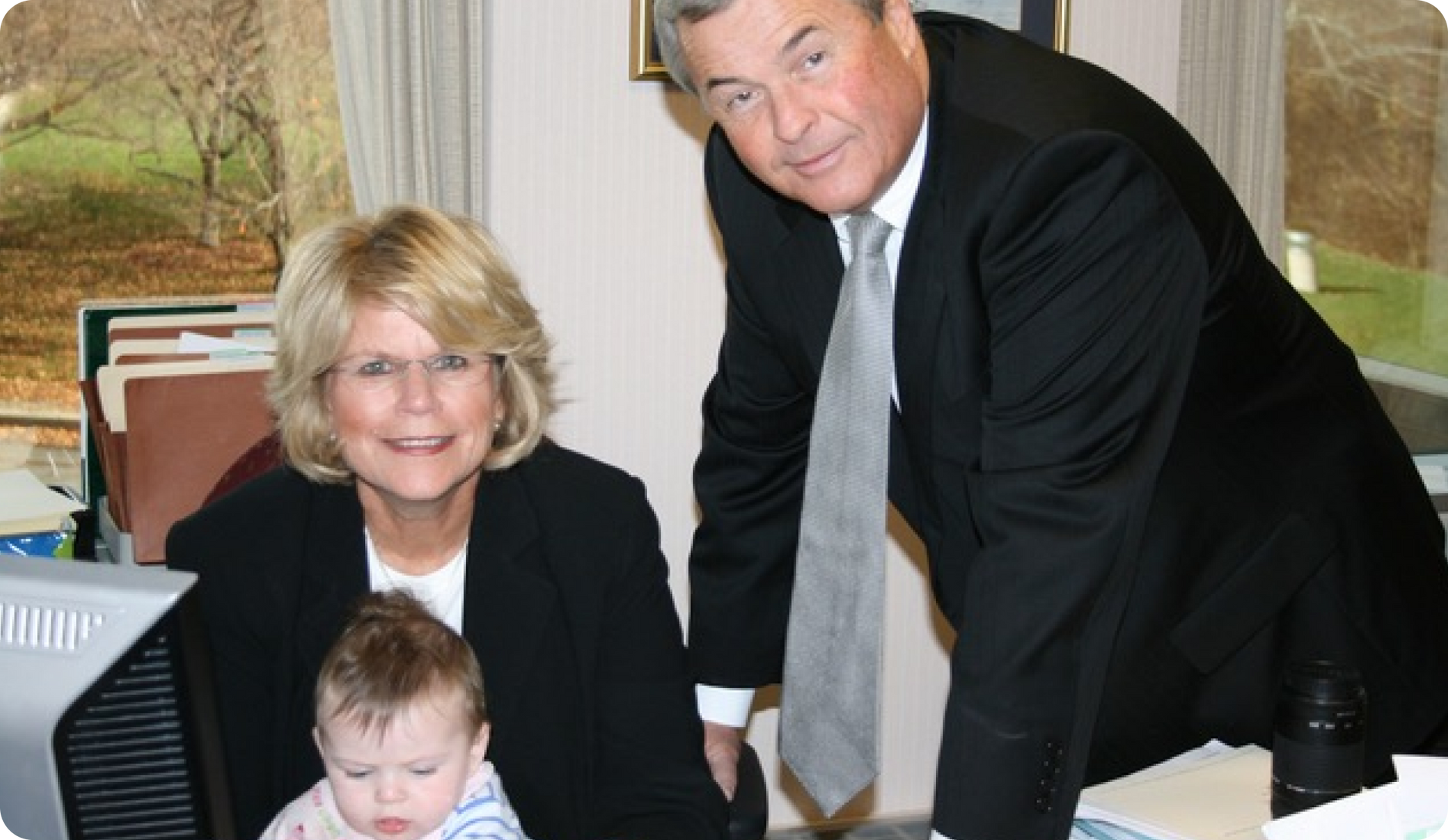 Woman and man smile, looking at camera with a baby. They are in an office setting.