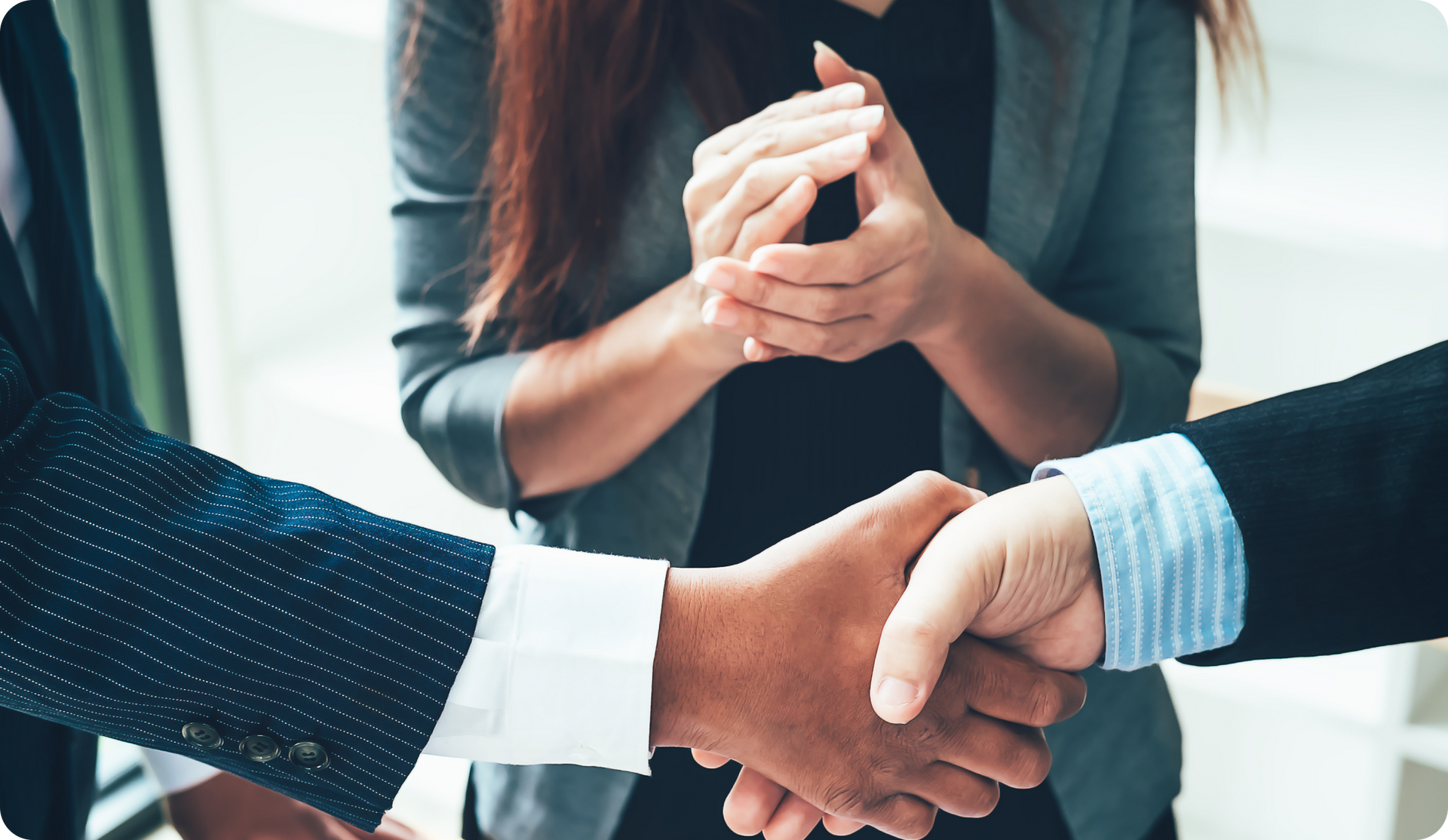 Two people in suits shaking hands, a woman in a suit clapping in the background.
