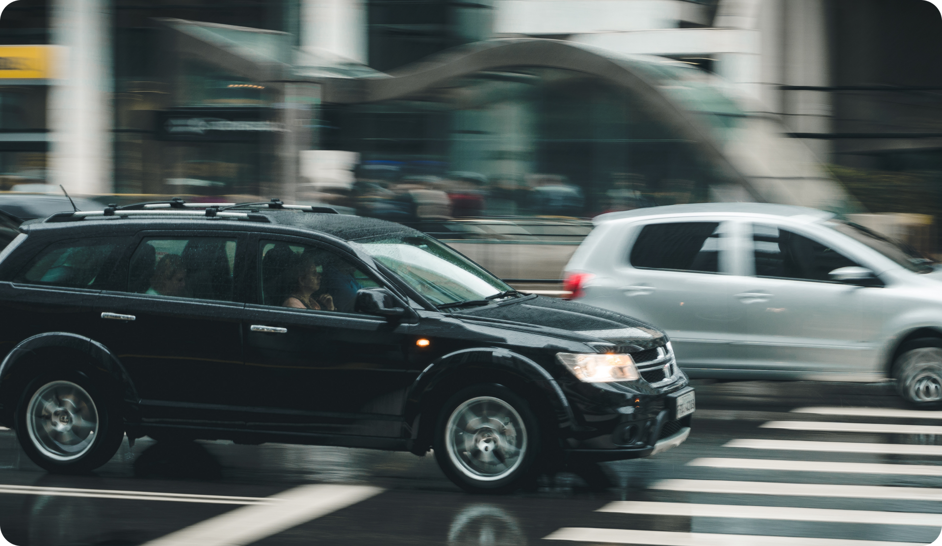 Black SUV drives across a wet street, blurred cityscape in the background; another car beside it.