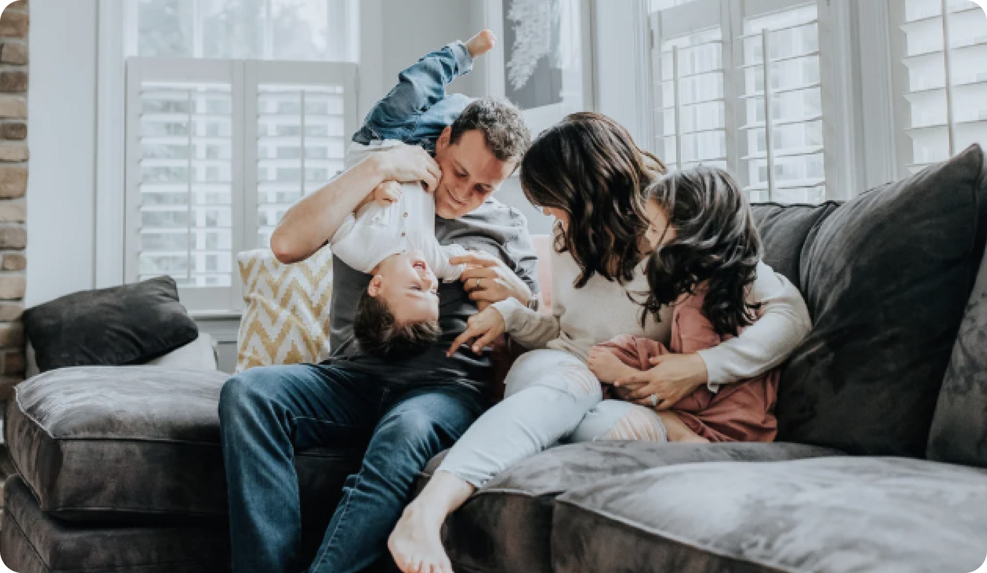 Family of four laughing on a gray couch. Father holds a child upside down, while mother and other child smile.