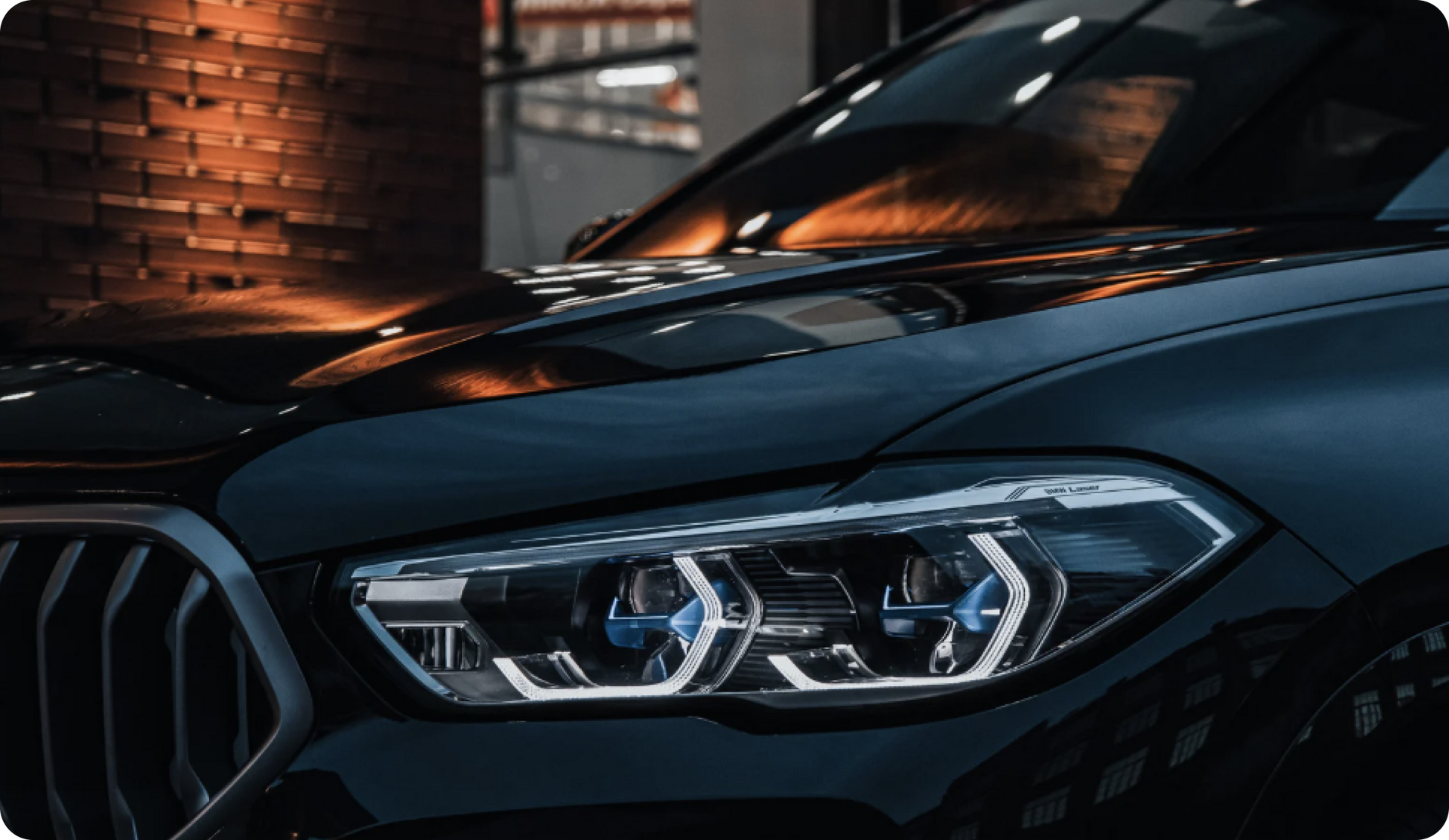 Black BMW SUV headlight detail, illuminated, parked, with brick wall background.