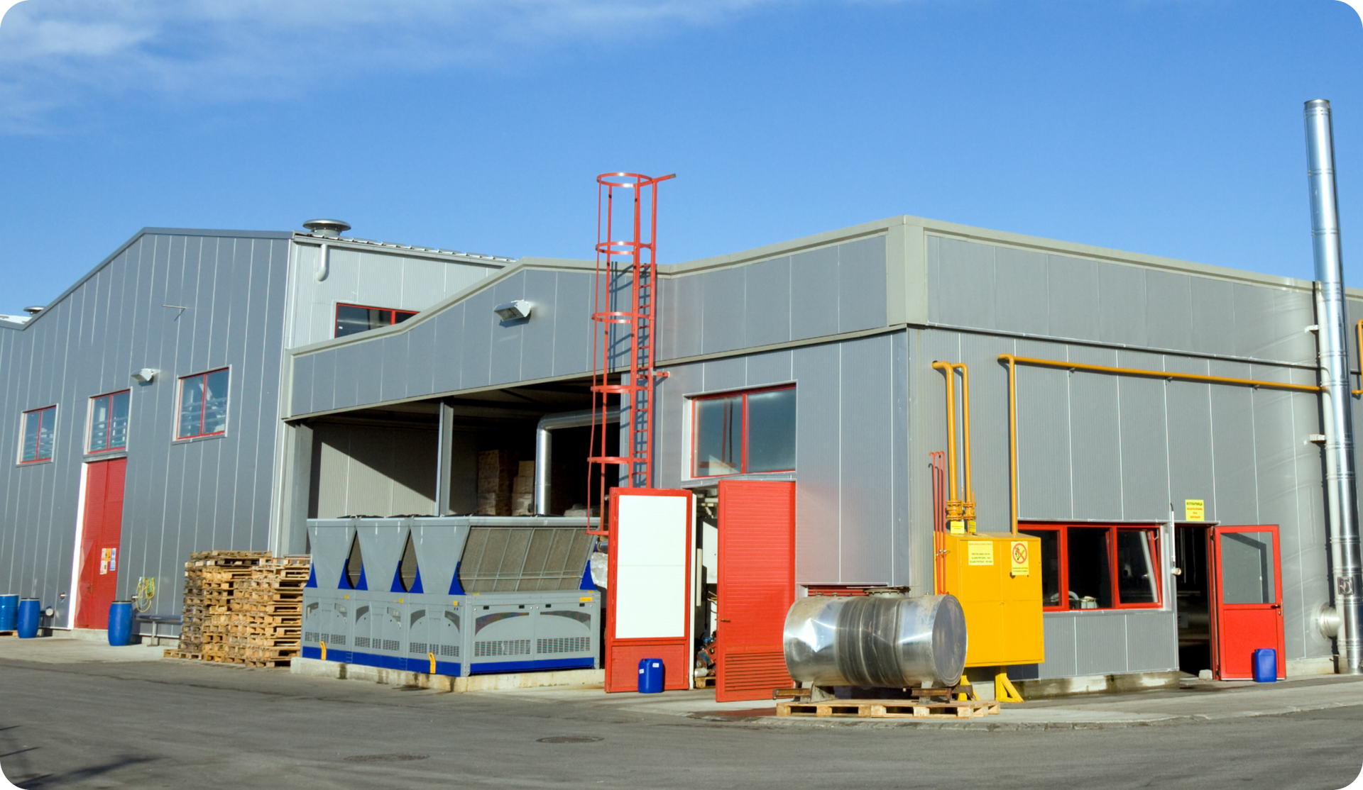 Exterior view of a factory building with gray metal siding, red doors, and various industrial equipment outside.