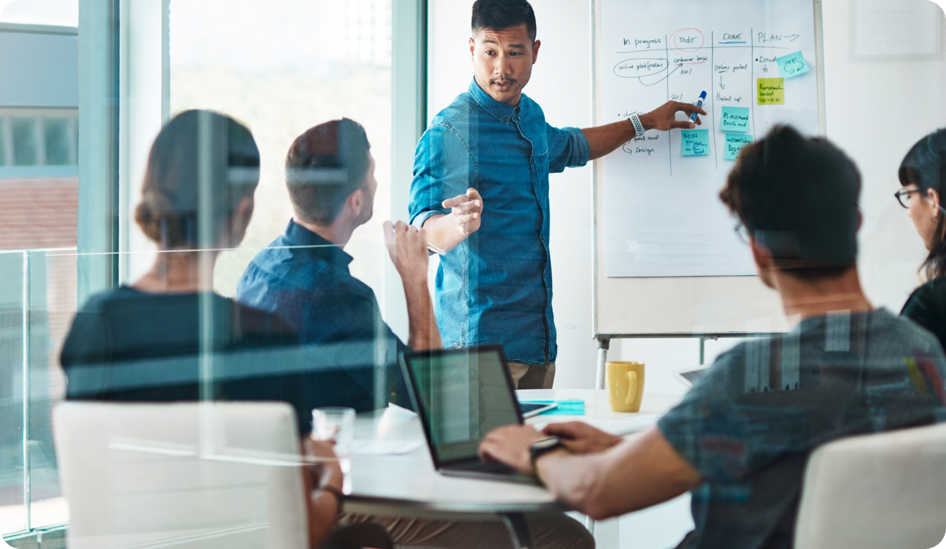 A man points to a whiteboard during a presentation to a group of people in a modern office.