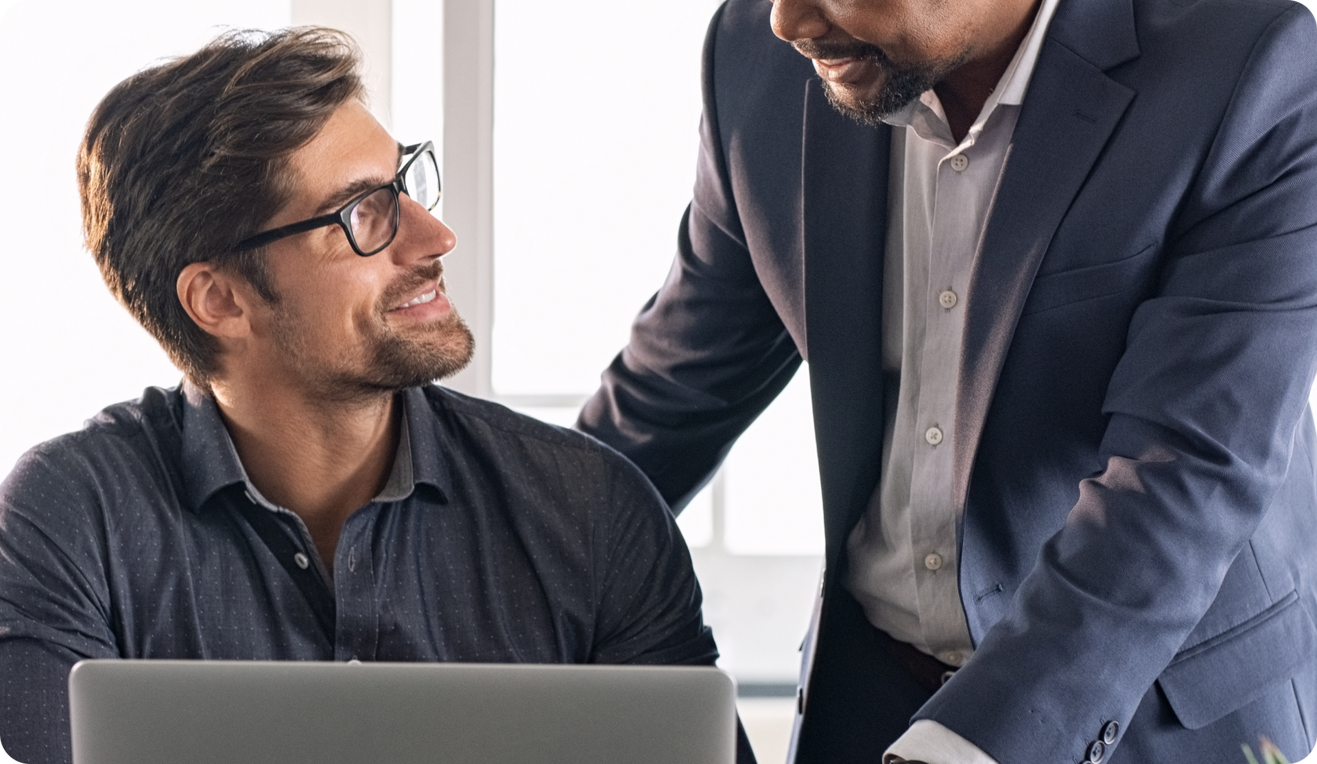 Two men at a desk, one in a suit and touching the other's shoulder, smiling. Office setting.