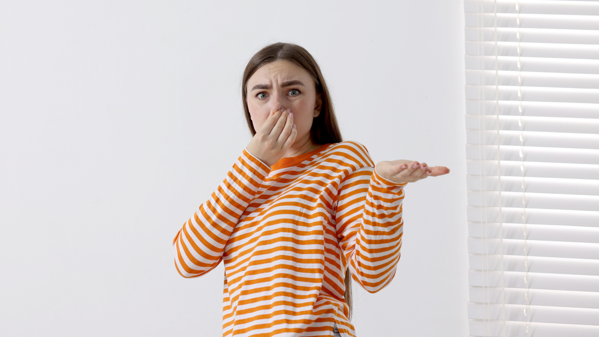 Woman covering her nose with a look of disgust, orange striped shirt, window with blinds.