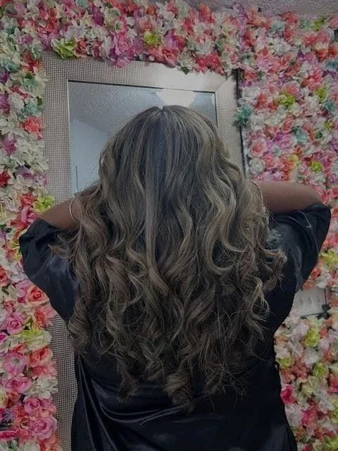 A woman with long curly hair is standing in front of a wall of flowers.