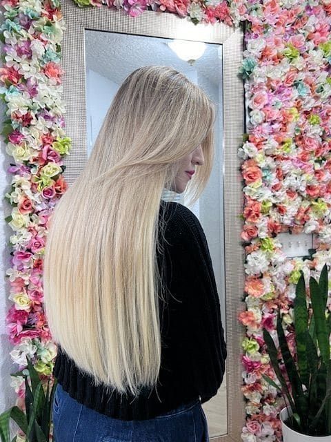 A woman with long blonde hair is standing in front of a mirror in front of a wall of flowers.
