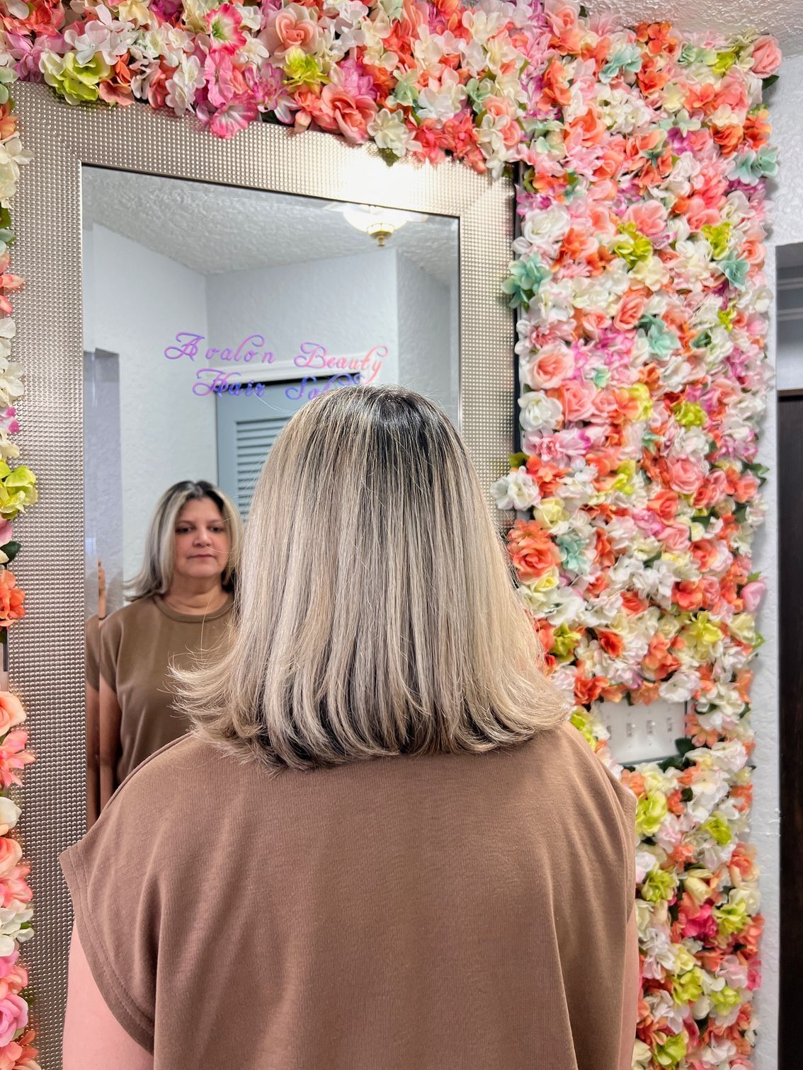 A woman is looking at her reflection in a mirror in front of a wall of flowers.