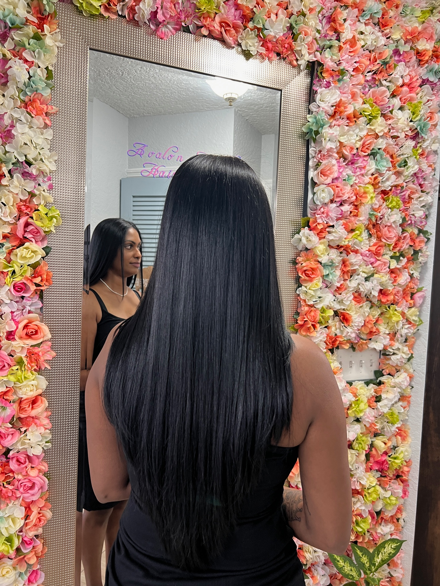 A woman is standing in front of a mirror with flowers on the wall behind her.