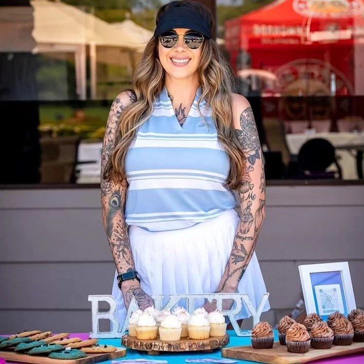 Woman with tattoos, sunglasses, and visor, smiling at bakery stand.