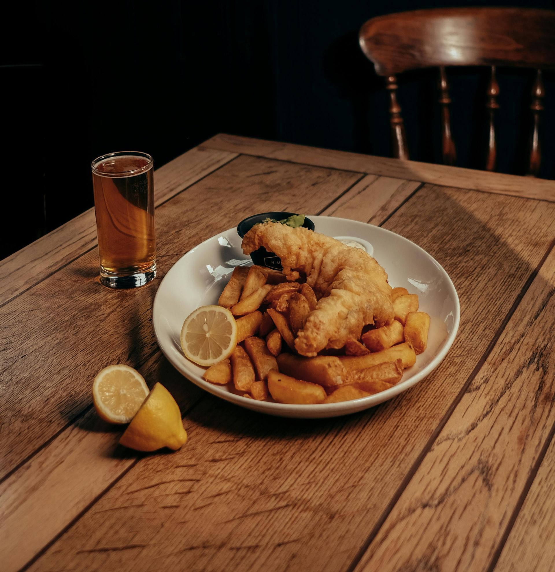 A plate of fish and chips on a wooden table next to a glass of beer