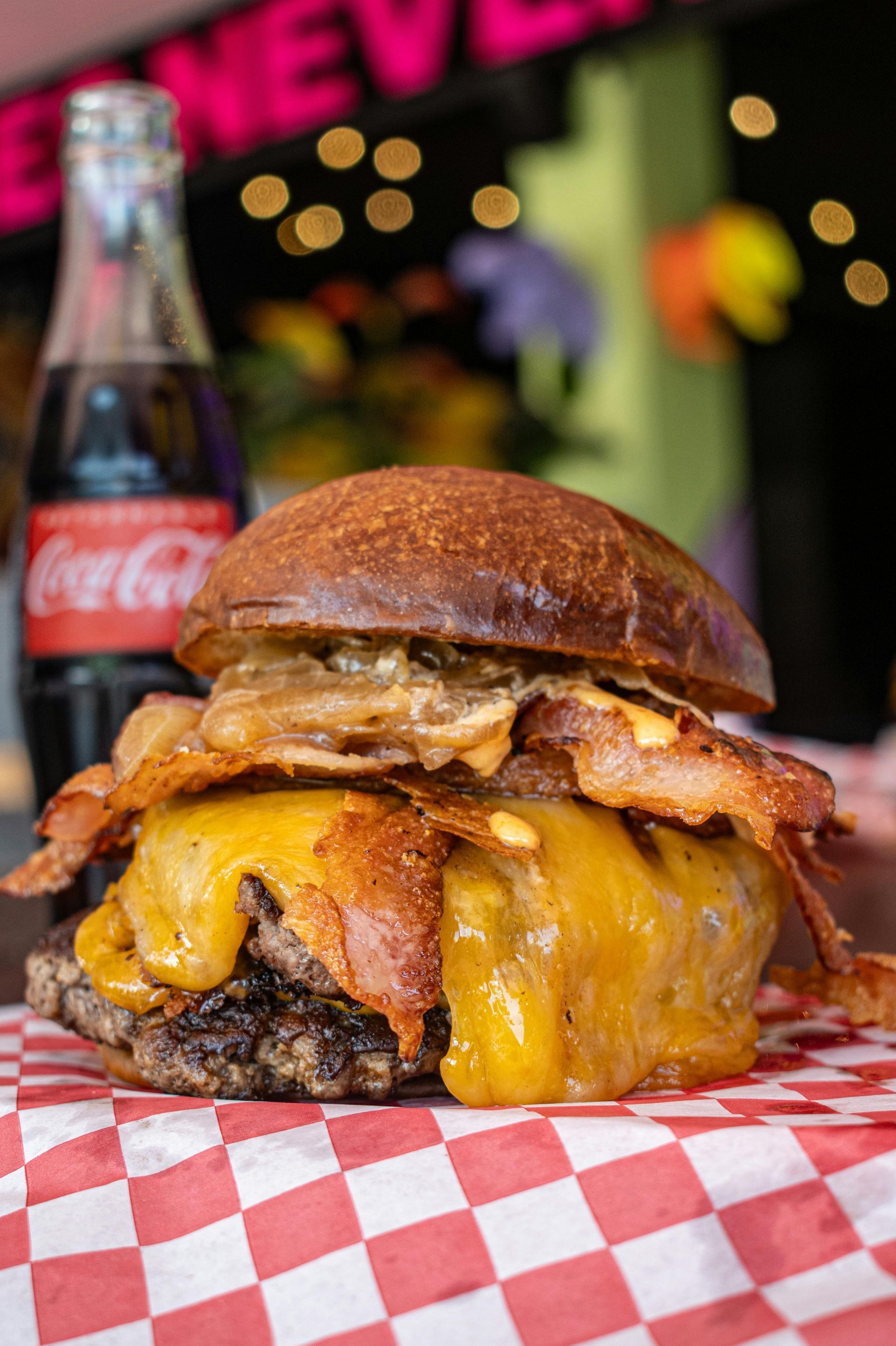 A hamburger with bacon and cheese is sitting on a checkered napkin next to a coca cola bottle.