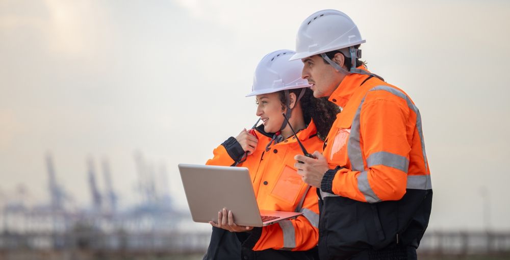 Two construction workers in orange vests and hard hats looking at a laptop at a construction site.