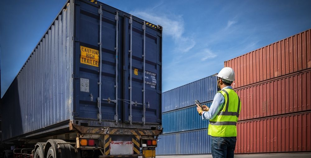 Person in safety vest and hard hat inspects cargo container at a port, holding a tablet.