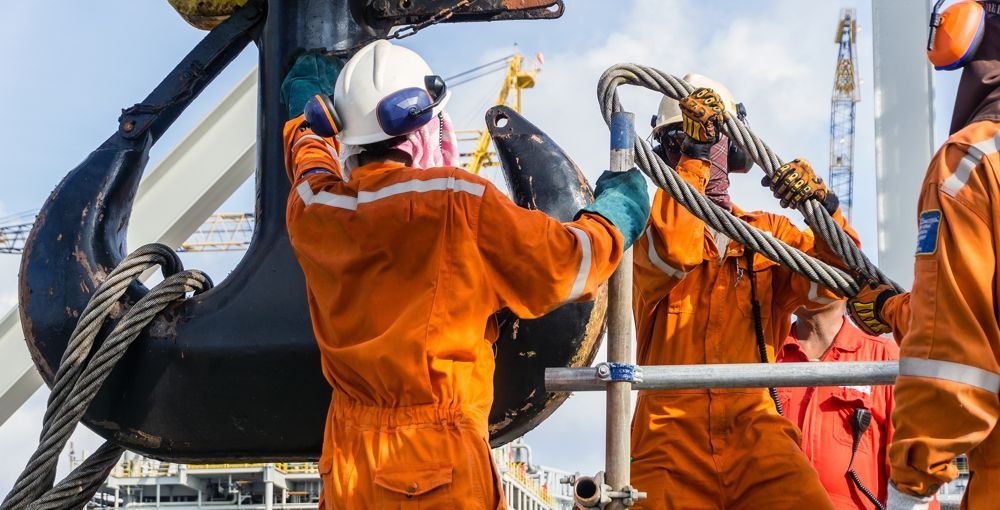 Workers in orange jumpsuits attaching a cable to a large black hook on a construction site.
