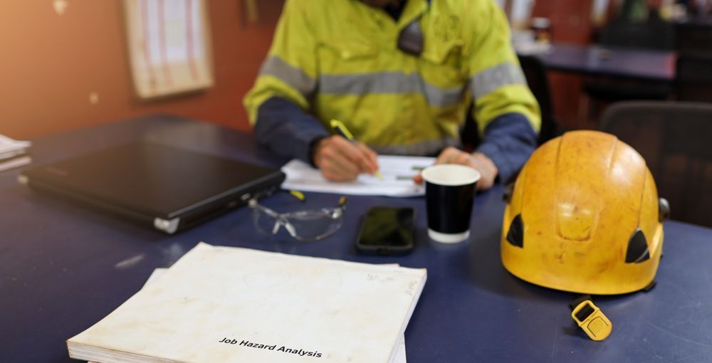 Construction worker writing at desk with hard hat, coffee, and papers.