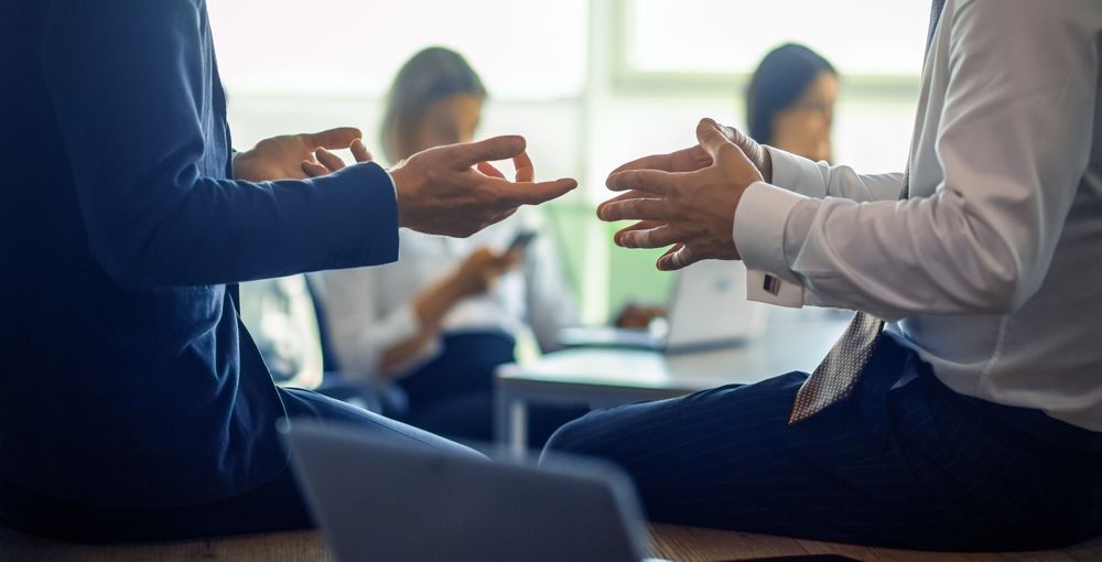 Two businesspeople gesturing with hands, sitting in front of a blurred office setting.