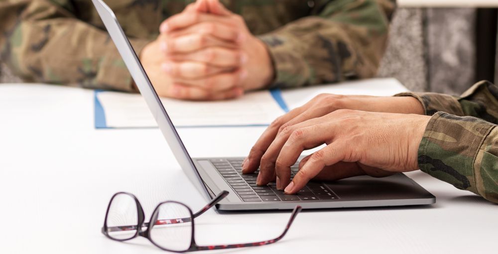 Two people in military uniforms at a table, one typing on a laptop, the other with hands clasped. Glasses in foreground.