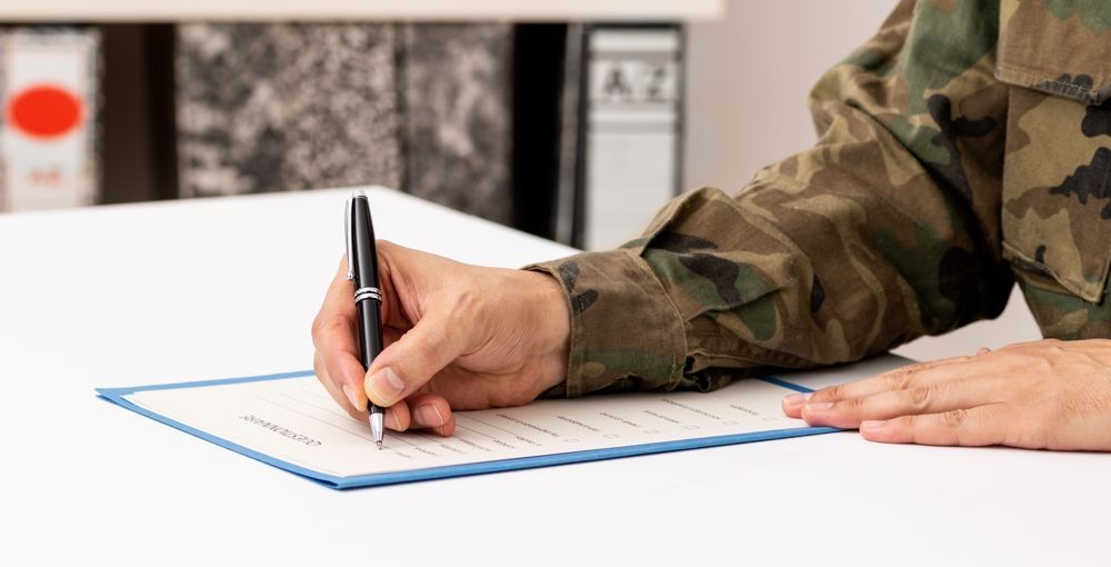Person in camouflage uniform writing on a document with a pen.