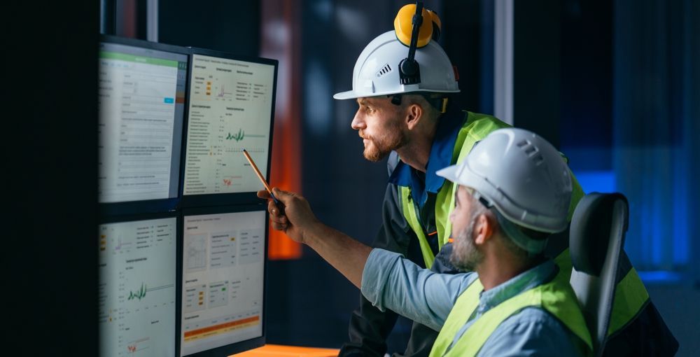Two engineers in hard hats examining data on computer monitors. One points.