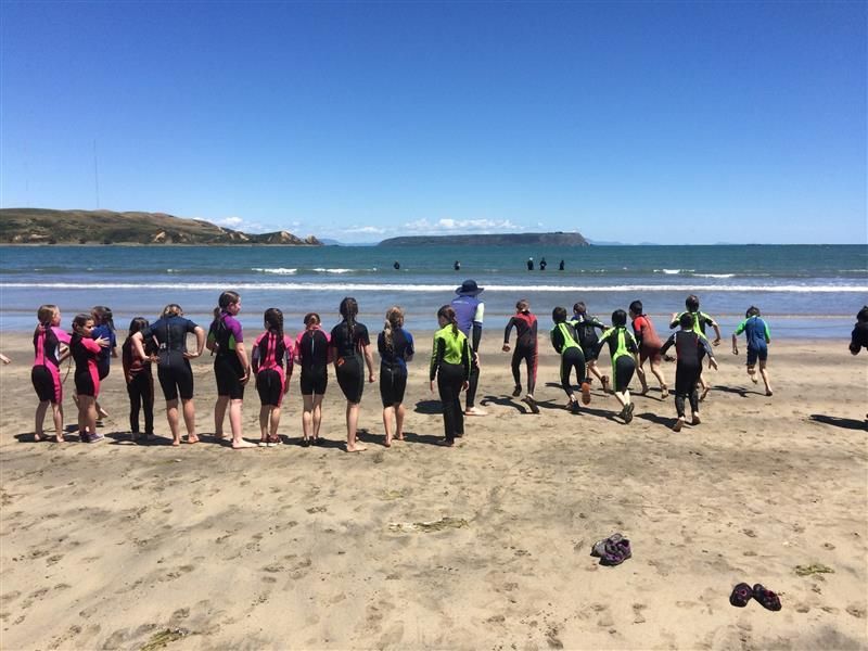 Kids in wetsuits on a beach, preparing to swim. Sunny day, blue sky, ocean in background.