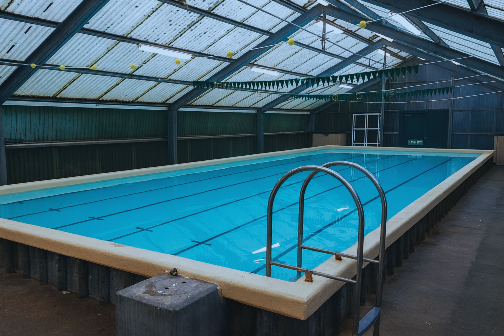 Indoor swimming pool with blue water and white borders under a corrugated metal roof.