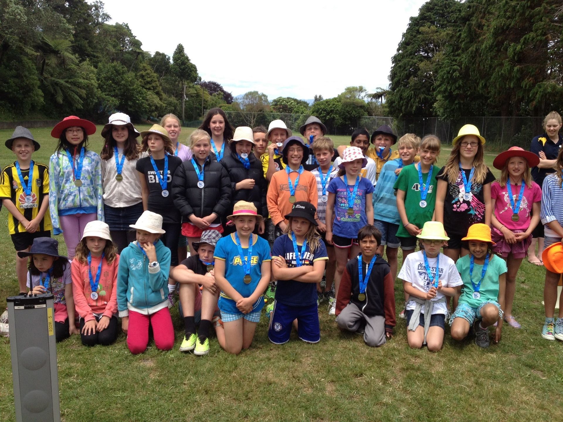 Group of children with medals on a grassy field, smiling, wearing hats, in a park.