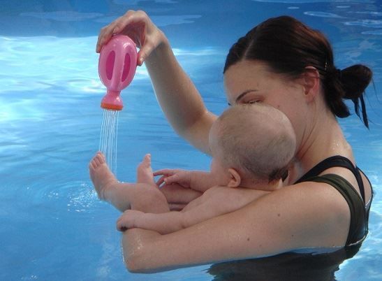Woman holding a baby in a pool, pouring water from a pink watering can.