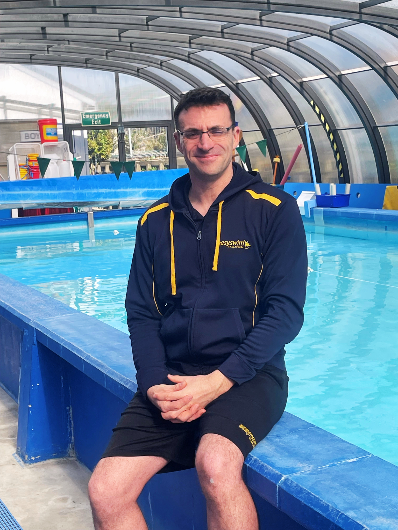 Man in navy jacket and shorts sitting by a pool, smiling, indoor pool setting.