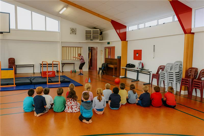 Children sit watching a teacher in a gym; equipment and chairs are visible.