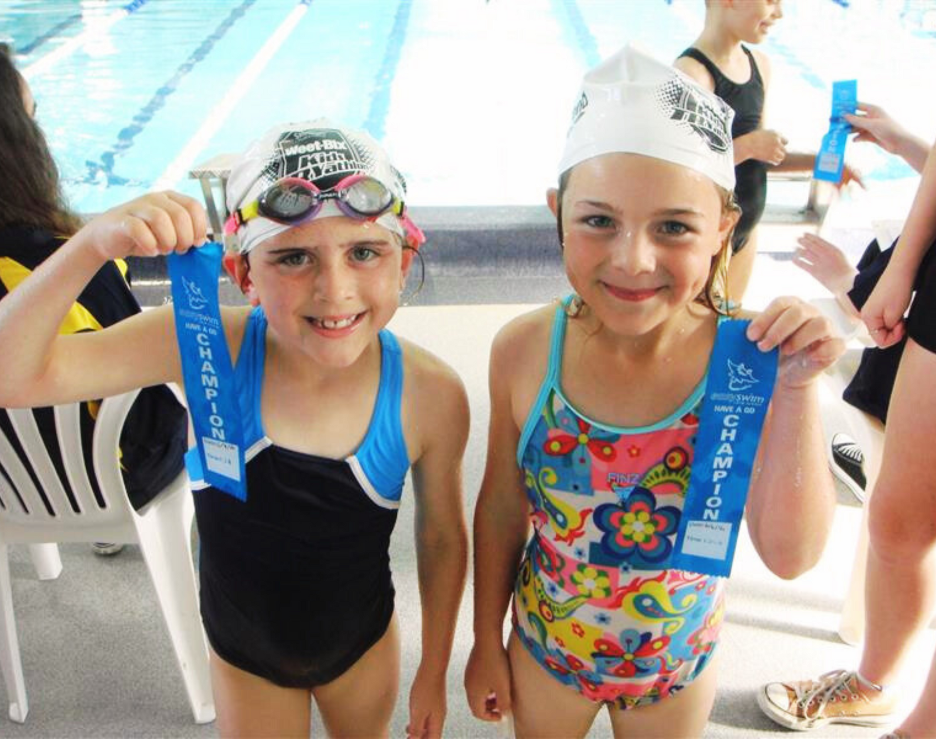 Two smiling girls in swimsuits hold blue ribbons by a pool. 