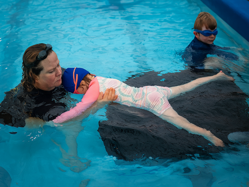 A swimming coach in pool, assisting child with swim instruction; boy in the background watches.