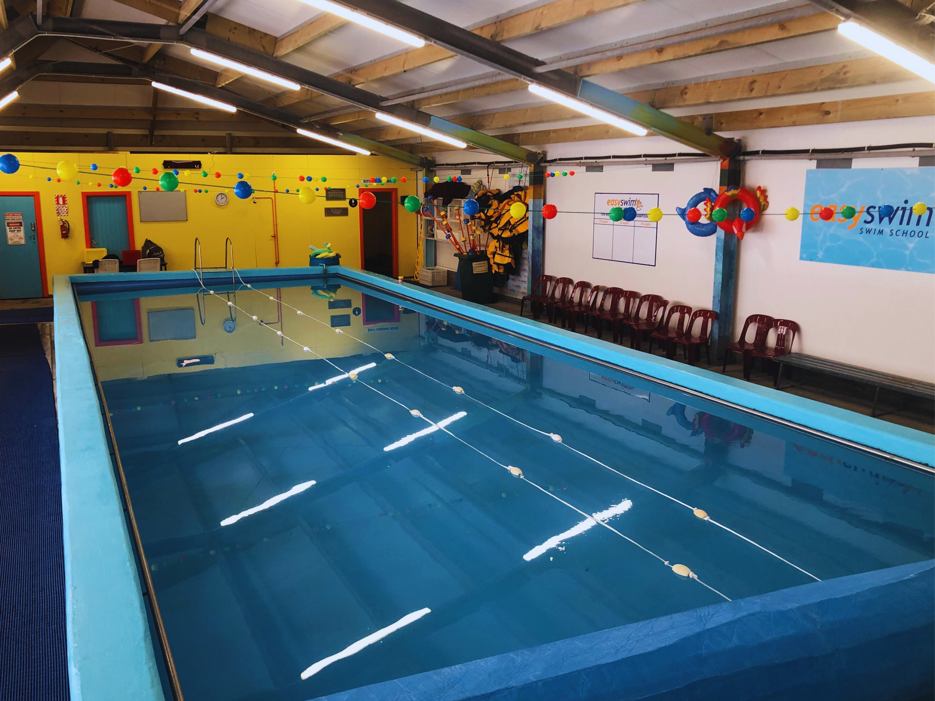 Indoor swimming pool, blue water, lane markers, yellow wall with balloons, blue pool edge, chairs.