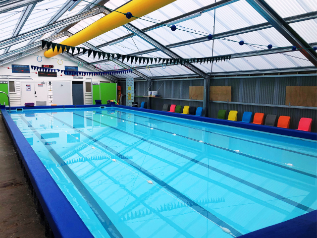 Indoor swimming pool with clear water and blue trim, under a glass roof. Colorful seating lines one side.