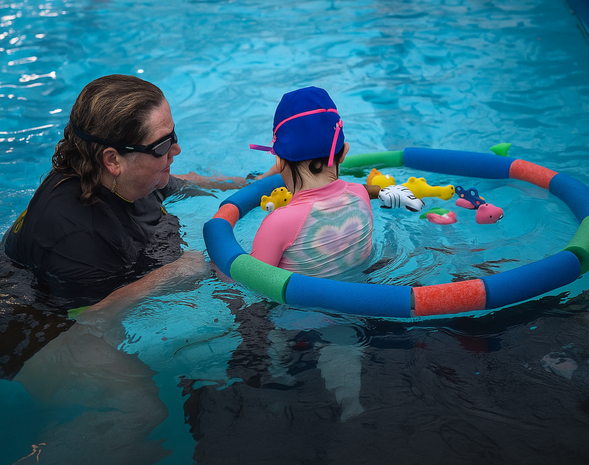 A swimming coach and a child in a pool; child wearing swim gear and in a floating ring with toys, the coach assists.