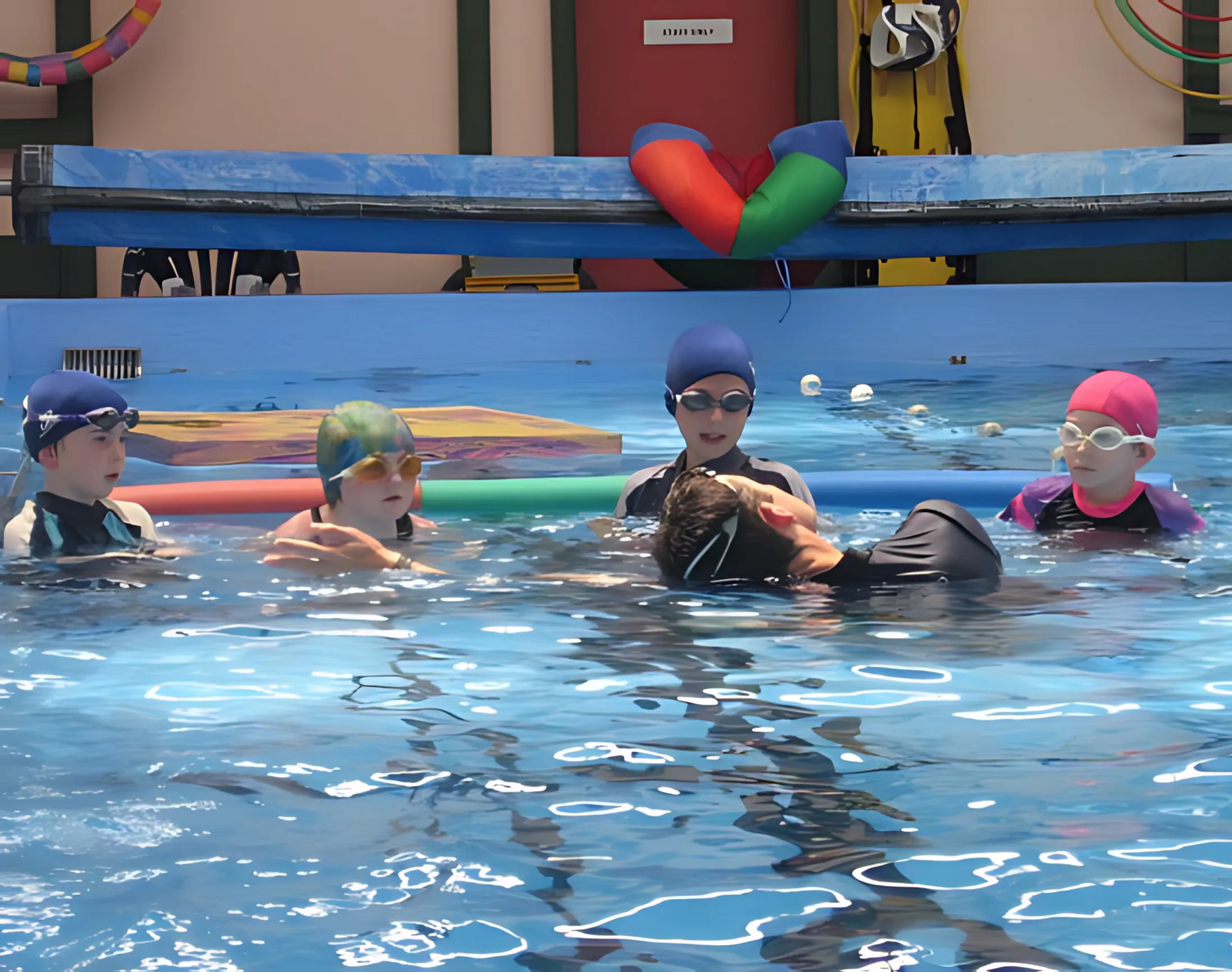 Children in a pool practicing swimming, with instructor helping. Blue water, colorful pool noodles.